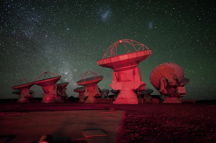 READY FOR USE. The Atacama Large Millimeter-submillimeter Array (ALMA) antennae bathed in red light. in the background there is the southern Milky Way on the left and the Magellanic Clouds at the top. ESO/C. Malin