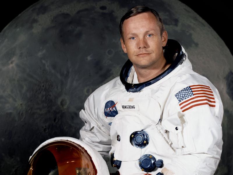 Portrait of Astronaut Neil A. Armstrong, commander of the Apollo 11 Lunar Landing mission in his space suit, with his helmet on the table in front of him. Behind him is a large photograph of the lunar surface. Image Credit: NASA