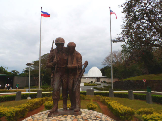 BROTHERS-IN-ARMS. The statue depicts two soldiers who have weathered the war. Behind them is the Pacific War Memorial 