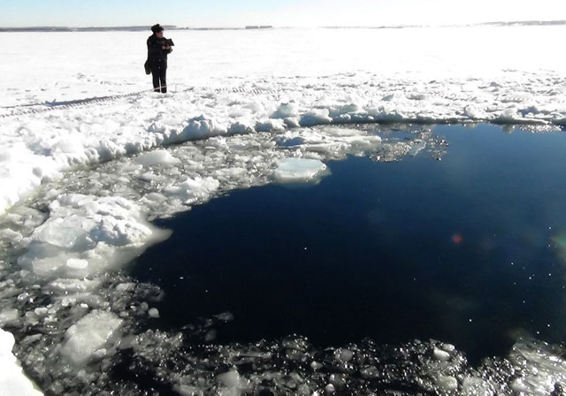 RUSSIAN FEDERATION, - : A handout photo taken on February 15, 2013, and provided by Chelyabinsk region police department shows a police officer standing near a six-metre (20-foot) hole in the ice of a frozen lake, reportedly the site of a meteor fall, outside the town of Chebakul in the Chelyabinsk region. Divers scoured today the bottom of a Russian lake for fragments of a meteorite that plunged to Earth in a blinding fireball whose shockwave injured 1,200 people and damaged thousands of homes. AFP PHOTO / CHELYABINSK REGION POLICE DEPARTMENT